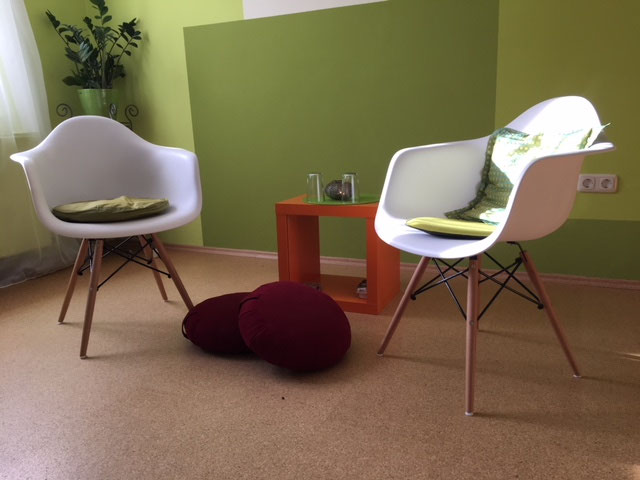 Zwei weiße Stühle mit farbigen Kissen stehen sich im Beratungsraum gegenüber, dazwischen ein orangefarbener Beistelltisch